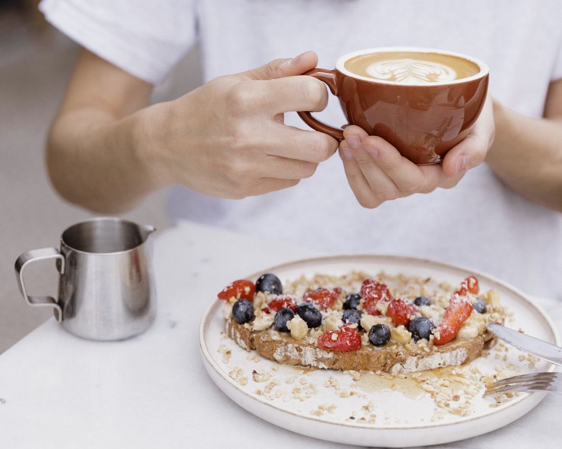 A woman holding a flat white next to a plate of toast with berries and granola at ARVO Bilbao